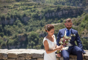 Plano medio de los novios, ella de pie y él sentado sobre un murete con paisaje de montaña de fondo. Reportaje de boda Almudena y Sergio por Andrea Aragón.
